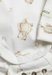 Une combinaison en coton avec des motifs de petit ours en peluche beige clair et de feuilles sur fond blanc. Présente une fermeture à pression et des poignets côtelés pour le confort.