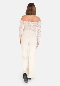 Body con in pizzo bianco con spalle scoperte, abbinato a pantaloni beige a gamba larga. Scarpe con tacco alto nere visibili da dietro.