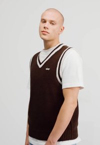 Vesta tricotată maronie cu accente albe peste un tricou alb; are un guler în formă de V și o textură cu nervuri. Vesta are un logo pe piept.