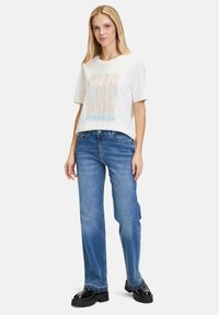 Hvid kortærmet t-shirt med "CIAO" grafisk design i multifarvede rhinsten, parret med blå lige ben jeans og sorte platform støvler.