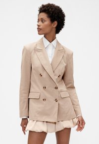 Bež dvoredno sako s velikim gumbima, oštrim reverima i bočnim džepovima, nošen preko bijele košulje i lagane široke suknje.