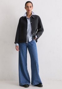 Czarna kurtka z mieszaniny wełny z kołnierzem, przednią kieszenią i zapięciem na guziki, noszona na niebieskiej koszuli w paski, w połączeniu ze spodniami o szerokich nogawkach.