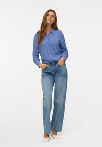 Blårandig skjorta med knappar och avslappnad passform, tillsammans med högmidjade ljusblå jeans. Modellen poserar med händerna i fickorna.