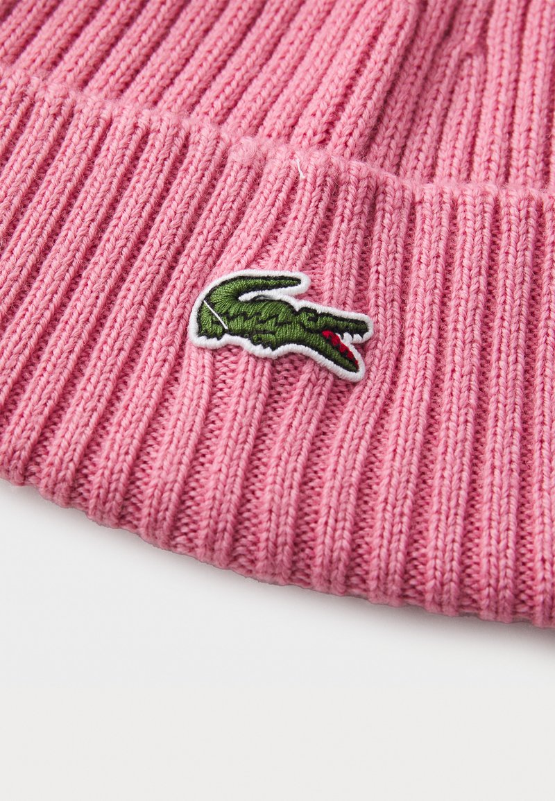 Różowa dzianinowa czapka z prążkowanej wełny z zielono-białym haftowanym logo krokodyla. Miękka tekstura, cylindryczny kształt, idealna do codziennego użytku.
