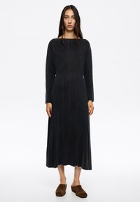 Vestido largo negro con escote ancho y mangas largas. Presenta costuras marrones en contraste y una silueta fluida. La modelo lleva mules de ante marrones.