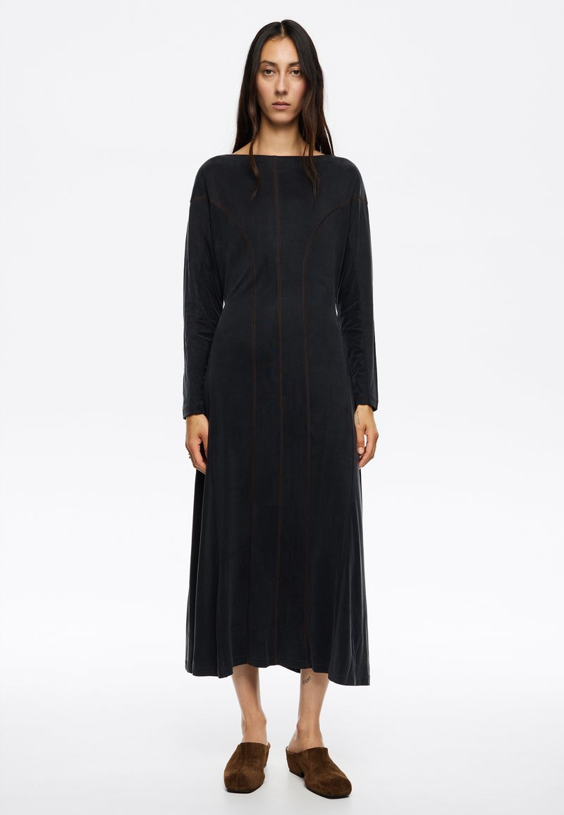 Vestido largo negro con escote ancho y mangas largas. Presenta costuras marrones en contraste y una silueta fluida. La modelo lleva mules de ante marrones.