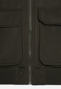 Alyvuogių žalio spalvos striukė su užtrauktuku priekyje, dviem didelėmis kišenėmis, raštuotu apačiu ir tekstūruotu audiniu. Yra sustiprintos siūlės ir matinė apdaila.