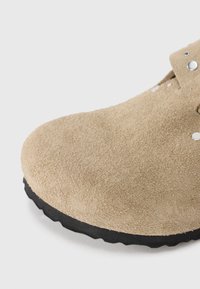 Gros plan sur une chaussure en daim beige avec une semelle texturée noire et des clous en métal sur la partie supérieure de la chaussure.