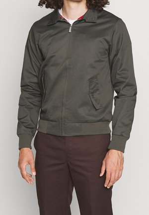 Homme portant une veste zippée vert olive avec poches à rabat, poignets et ourlet côtelés, associée à un pantalon marron foncé, debout sur un fond uni.