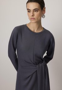 Vestido gris oscuro hecho de tela suave, con mangas largas, escote redondeado y un detalle retorcido en la cintura para dar forma adicional.