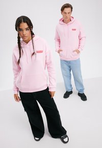 Hoodie rose clair avec le logo "DELICIOUS", poche avant, associé à un pantalon noir ample et des baskets noires. Le mannequin pose sur un fond clair.
