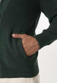 Tumedast roheline kudumitega kardigan ribatekstuuriga, millel on esitasku ja ribakäised. Materjal on paks ja soe.