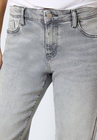 Close-up de jeans de denim cinza claro com botão frontal, passadores de cinto e bolsos, usados por uma pessoa com uma blusa branca.