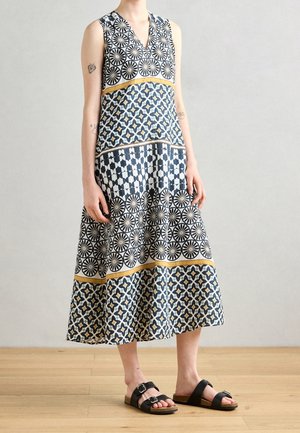 Ärmelloses Kleid mit geometrischen Mustern in Schwarz, Weiß und Gelb, getragen mit schwarzen Sandalen mit Schnalle vor einem Holzboden.
