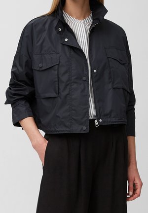 Persoon die een zwart cropped utility jack met drukknopen zakken en een gestreept hemd eronder draagt, hand in zak, staand tegen een effen achtergrond.