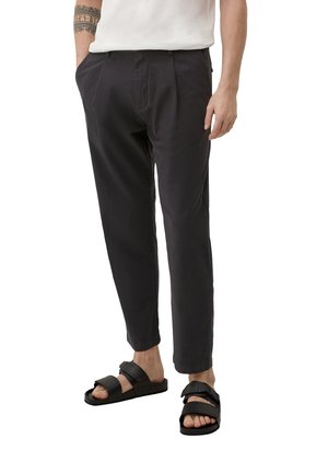 Mand iført sorte tilspidsede bukser og sorte sandaler med to remme, står med den ene hånd i lommen mod hvid baggrund.