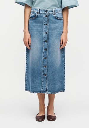 Γυναίκα που φοράει φούστα από τζιν με κουμπιά στο μπροστινό μέρος μέχρι τη μέση, ανοιχτό μπλε μπλούζα και σκούρα καφέ επίπεδα παπούτσια, στέκεται με τα χέρια στα πλευρά.