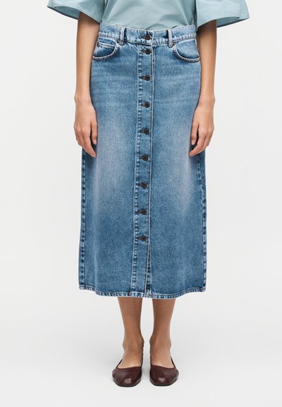 Femme portant une jupe en jean mi-longue boutonnée sur le devant, un chemisier bleu clair et des chaussures plates marron foncé, debout les bras le long du corps.
