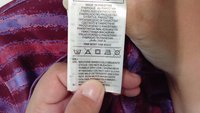 Textile care label held in hand, white fabric with black printed text, includes washing symbols, instructions, and multilingual information.