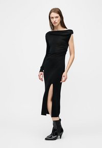 Femme portant une robe noire asymétrique à manches longues avec une fente haute sur la cuisse et des bottines noires pointues, posant devant un fond blanc.