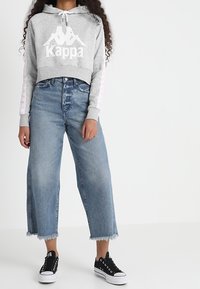 Felpa grigia corta con logo Kappa bianco e dettagli rosa sulle maniche. Abbinata a jeans azzurri a gamba larga e sneakers nere.