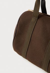 Καφέ τσάντα duffle από τσόχα με σκούρες ελαιόλαδες υφασμάτινες λαβές και χρυσό φερμουάρ σε λευκό φόντο.