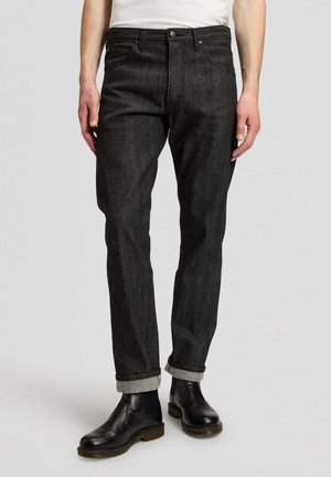 Mann trägt schwarze hochgekrempelte Jeans und schwarze Lederankleboots und steht vor einem schlichten Hintergrund.