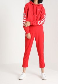 Felpa rossa con cappuccio e maniche lunghe con scritte bianche, abbinata a pantaloni jogging rossi coordinati e sneakers bianche. Tessuto liscio, vestibilità comoda.
