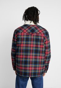 Rutigt skjorta med röda, blåa och gula mönster, med en mjuk, vit pälskrage och knäppningar på ärmarna.