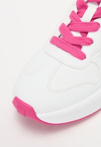 Sneaker blanche avec des accents roses, en matériau synthétique texturé, à bout rond, semelle plate et lacets roses. Détail de couture notable le long des côtés.