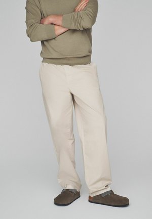 Beige Chinos mit lockerem Sitz, mit glatter Oberfläche und Seitentaschen. Kombiniert mit dunkelbraunen Slippern. Hellgrauer Hintergrund.