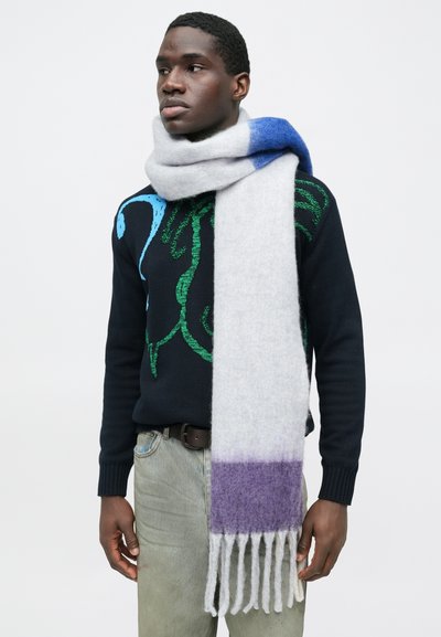 Άνδρας που φοράει σκούτερο πουλόβερ με πράσινα και μπλε σχέδια, ανοιχτόχρωμο τζιν και ένα χοντρό λευκό κασκόλ με μπλε και μοβ χρωματικές μπλοκ.