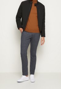 Zwarte gewatteerde jas, bruin trui, grijze slim-fit broek en witte sneakers. Jas heeft een opstaande kraag; broek heeft een zachte textuur.