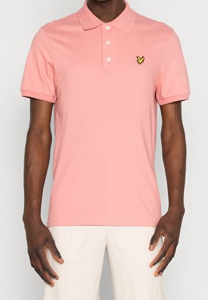 Mand iført en lyserød kortærmet polo-shirt med tre knapper og et lille gult fugle-logo på venstre bryst.