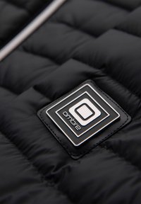 Tissu matelassé noir avec un patch de marque carré cousu présentant un motif géométrique et le mot "ombre" en lettres minuscules.