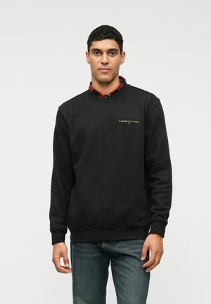 Sweatshirt preto com decote redondo, punhos e barra canelados. Apresenta um logotipo frontal em amarelo. Usado sobre uma camisa xadrez vermelha.