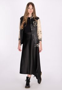 Zwarte en beige varsity jack met crèmekleurige mouwen, zwarte satijnen rok en zwarte veterschoenen. Lange haren, staand tegen een witte achtergrond.
