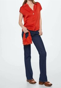 Chemise à boutons rouge à manches courtes, jeans évasés bleu foncé, sabots en plateforme marron et un petit sac à franges orange.
