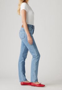 Vaqueros de mezclilla azul con pernera recta, cintura alta y una etiqueta de cuero marrón en la parte trasera. Combinados con una camiseta blanca ajustada y bailarinas rojas.