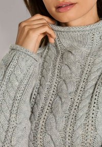 Nahaufnahme einer Person, die den Kragen eines hellgrauen, grob gestrickten Sweaters mit geflochtenen Mustern an Ärmeln und Körper anpasst.
