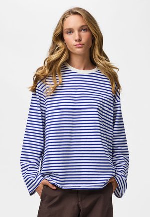 Jeune femme aux longs cheveux blonds portant une chemise à manches longues rayée bleue et blanche et un pantalon marron, mains dans les poches, fond neutre.