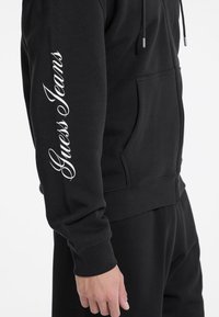 Sort zip-up hoodie med hvid "Guess Jeans" tekst på venstre ærme, lavet af blød bomuld, med en frontlomme og justerbare snore.