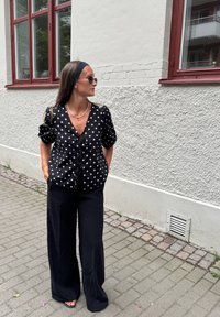 Kvinna med långt hår i svarta solglasögon, svart blommig blus och vida svarta byxor står på en platt trottoar mot en vit vägg.