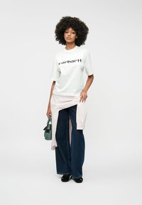 Fehér póló fekete logóval, egy világos rózsaszín inggel rétegezve, laza szabású kék farmernadrággal és fekete cipővel, zöld táskával a kezében.