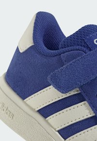 Modré semišové tenisky s bílými koženými pruhy, suchým zipem, texturovanou tkaninou a gumovou podrážkou. Doplňky zahrnují prošívané logo.