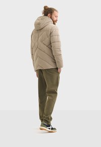 Chaqueta acolchada beige con capucha, con costuras diagonales. Combinada con pantalones verdes oliva y zapatillas negras con acentos blancos y suela color goma.