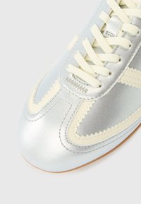 Sapatilha de couro prateado com detalhes em creme contrastantes, biqueira redonda, costura texturizada e atacadores planos. Design leve e elegante.