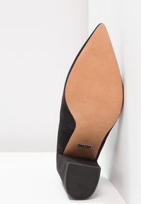 Chaussure noire en daim à bout pointu avec semelle en cuir couleur beige et talon bloc, taille 37, positionnée contre une surface d'angle blanche.