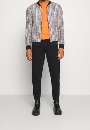 Geruite bomberjack in rood, zwart en wit; oranje T-shirt eronder; zwarte broek; zwarte enkellaarsjes. Casual pasvorm en ribbelmanchetten.
