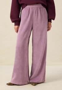 Pantalons larges en tissu lilas côtelé et doux. Présentent une taille élastique et une silhouette fluide, associés à des chaussures à motifs sombres.